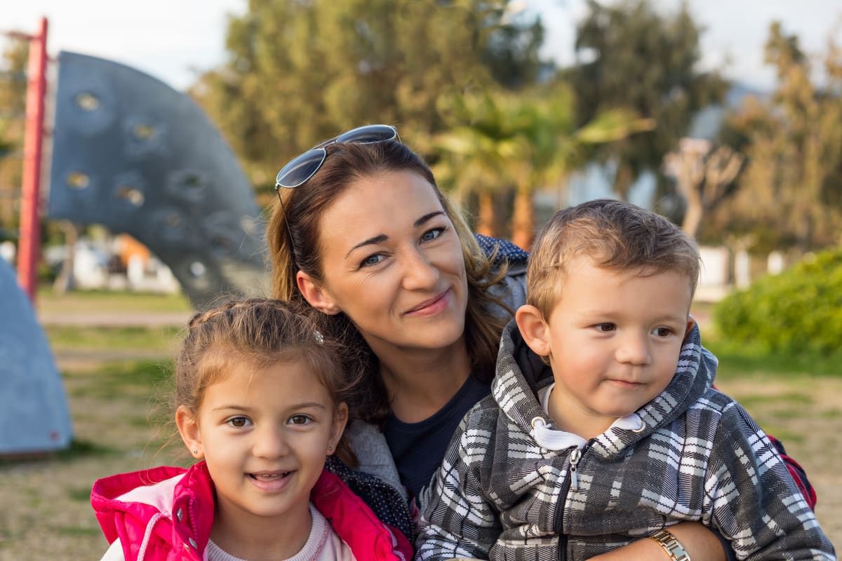 A mother smiling with her young daughter and son at a park playground